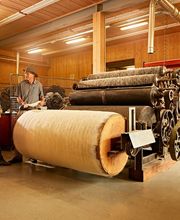 Drei Personen betrachten in einer Werkstatt eine große alte Textilmaschine mit Wollbahnen.