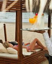 Persona sirviendo bebidas en la playa bajo una palapa, con dos personas relajándose en un sofá.