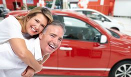 Smiling couple in front of a red car, with the woman playfully on the man's back.