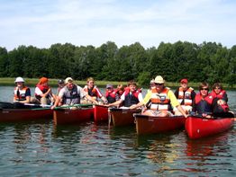 Eine Gruppe von Personen in Kanus auf einem See, alle tragen Schwimmwesten und genießen den sonnigen Tag.