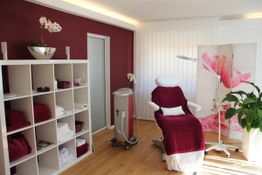 Kosmetiksalon mit roter Wand, weißem Regal, einem Behandlungsstuhl und Pflanzen im Raum.