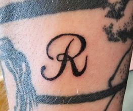 De letter "R" in cursief getatoeëerd op een arm met andere tatoeages eromheen.