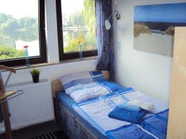 Kleines Schlafzimmer mit Bett, Fensterblick auf See, blau-weiße Bettwäsche, Pflanzen und Bild.