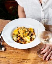 Pessoa à mesa com prato de massa e taça de vinho branco, vestindo camisa branca.