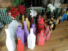 Garrafas coloridas decorativas em uma mesa de madeira, com flores vermelhas e brancas ao fundo.