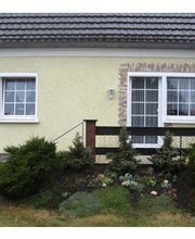 Kleines gelbes Haus mit Satteldach, zwei Fenstern und einem kleinen Vorgarten mit Sträuchern.