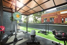 Fitnessbereich im Freien mit Gewichten und Bank, umgeben von Bäumen und einem Backsteingebäude.