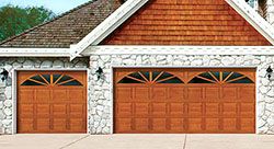 Two wooden garage doors with decorative windows on a stone and brick house facade.