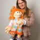 Eine Frau hält eine große Handpuppe mit rotem Haar. Die Puppe trägt ein buntes Blumenkleid.