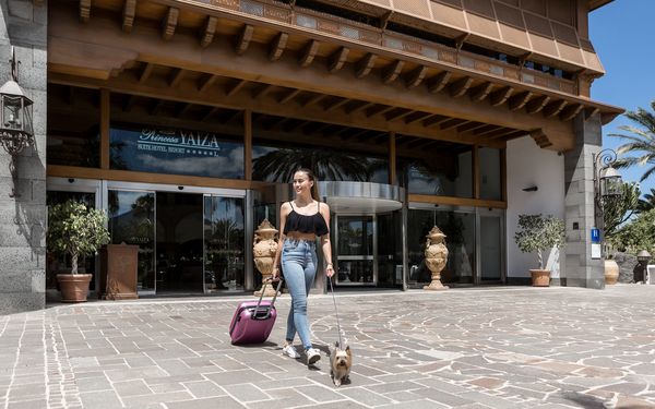 lächelnde Frau mit Hund an der Leine und einem Koffer in der Hand verlässt Hotel