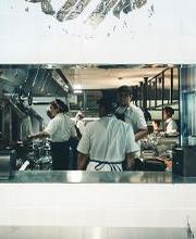 Cozinheiros trabalham em uma cozinha movimentada, vistos através de uma janela interna.