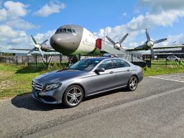 Grijze auto geparkeerd voor een oude propellervliegtuig op een zonnige dag met blauwe lucht.