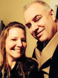 Man and woman smiling close together, indoors, man in tan coat, warm lighting.