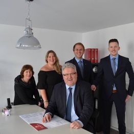 Fünf Personen in Business-Kleidung posieren in einem Büro, ein Mann sitzt am Tisch.