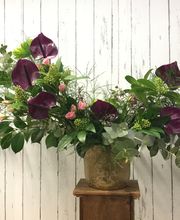 Arreglo floral en jarrón con flores moradas y rosas, sobre fondo de madera blanca.