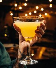 Mão segurando um copo de coquetel amarelo com borda açucarada, luzes ao fundo.