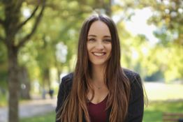 Junge Frau mit langen Haaren lächelt im Park unter Bäumen, sonniger Tag.
