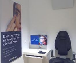 Kamer met een oogmeetstoel, poster over contactlenzen en een computermonitor met een gezicht.