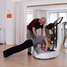 Eine Frau macht eine Plank-Übung auf einem Gerät, während ein Mann ihr Anweisungen gibt.
