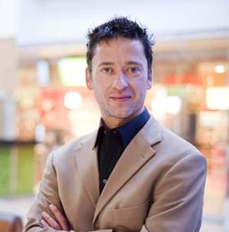 Mann in beigem Anzug mit verschränkten Armen, lächelt und steht vor unscharfem Hintergrund.