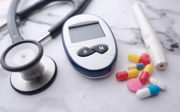 Ein Blutzuckermessgerät liegt auf einer weißen Marmorplatte, zwischen Diabetesmedikamenten und einem Stethoskop.
