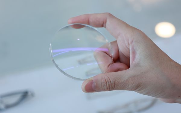 Eine Hand hält ein geschliffenes Brillenglas, vor einen neutralen weißen Hintergrund, ins Bild.