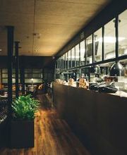 Restaurante moderno com cozinha aberta, piso de madeira e plantas decorativas.