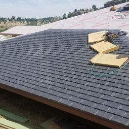 A roof in progress with gray shingles and construction materials on top, against a forested backdrop.