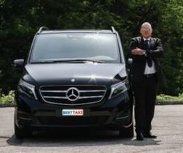Ein Mann steht in Anzug neben einem schwarzen Mercedes-Großraumwagen auf einer Straße.