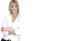 Frau in weißem Blazer steht vor weißem Hintergrund mit verschränkten Armen.