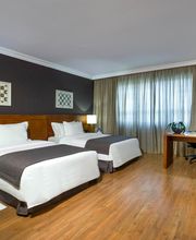 Quarto de hotel com duas camas, mesa, TV e janelas com cortinas num ambiente aconchegante.