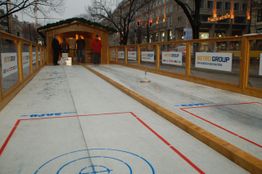 Zwei Eisstockbahnen mit Holzgeländer, Menschen im Hintergrund, Winterstimmung in der Stadt.