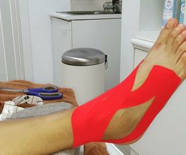 Pie con cinta kinesiológica roja en un consultorio. Tijeras y material médico al fondo.