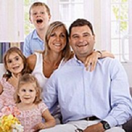 A smiling family of five posing together in a bright room with flowers on a table.