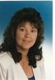 Eine Frau lächelt vor blauem Hintergrund. Sie trägt eine weiße Jacke und ein Kettenanhänger-Kreuz.