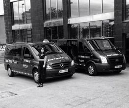 Dos taxis Mercedes Vito y Ford Transit estacionados frente a un edificio; una persona al lado izquierdo.