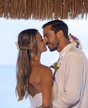 Pareja abrazada bajo techo de palma, frente al mar, intercambiando un beso.