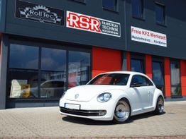 Weißer Volkswagen Beetle vor einer Werkstatt mit roten und schwarzen Fassaden.