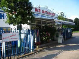 Imbiss "Der Sattmacher" mit blauer Gitterzaun, Sonnenschutz und Schild: "Bester Imbiss vor der Autobahn".