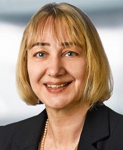Eine lächelnde Frau mit blondem Haar und einer schwarzen Jacke vor unscharfem Hintergrund.