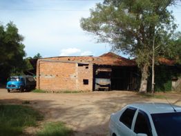 Galpão de tijolos em área rural com caminhões e carros estacionados ao redor, árvores ao fundo.