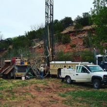 Drilling equipment and trucks set up near a rocky hillside and greenery.