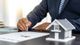 Person signing a document with a model house on the table.