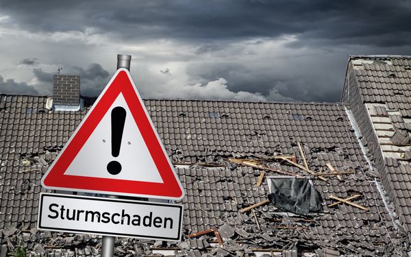 Vor einem zerstörten Dach steht ein Warnschild mit dem Hinweis auf einen Sturmschaden.