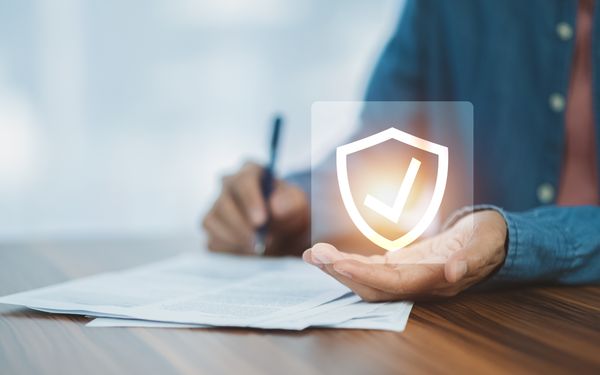 Ein Arbeitnehmer unterschreibt einen Versicherungsschutz mit der rechten Hand und hält in der Linken eine symbolische Grafik in der Hand, die ein Schutzschild und einen Haken darstellt.