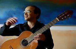 Mann mit Brille hält Gitarre, lächelnd vor buntem Hintergrund.