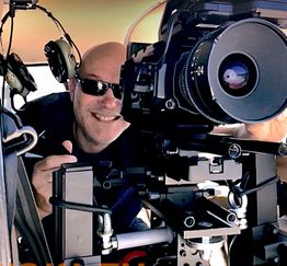 Eine Person mit Sonnenbrille bedient eine große Kamera mit Kopfhörer im Hintergrund.