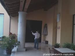 Hombre bajo un techo en patio con plantas y columnas, apuntando hacia una puerta oscura.