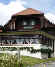 Altes Gasthaus mit Holzfassade und roten Dachziegeln, umgeben von grüner Landschaft, Himmel bewölkt.