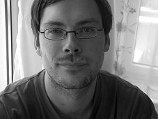 Ein Mann mit Brille und Bart schaut lächelnd in die Kamera, im Hintergrund ein Vorhang.